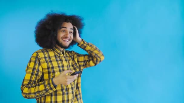 Feliz joven y atractivo afroamericano usando el teléfono y recibiendo buenas noticias sobre el fondo azul. Concepto de emociones — Vídeos de Stock