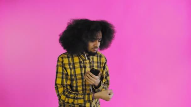 Uomo afroamericano positivo che usa il telefono per ascoltare musica nelle cuffie su sfondo viola. Concetto di emozioni — Video Stock