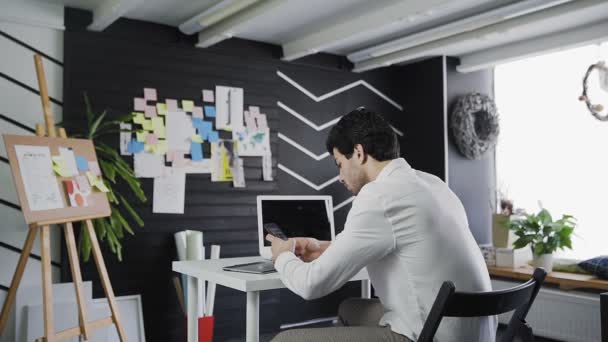 Młody atrakcyjny mężczyzna rozmawia, nagrywa i pisze na smartfonie w miejskim biurze, siedząc przy stole — Wideo stockowe