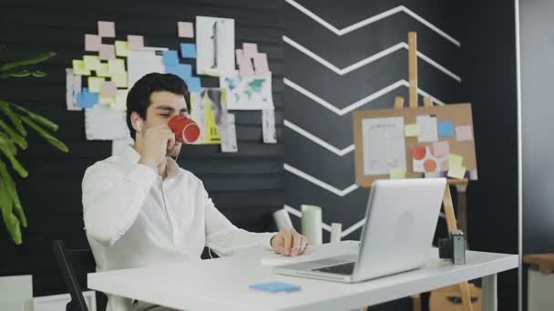 Glücklicher Geschäftsmann trägt Headset, spricht mit Blick auf Laptop und trinkt Kaffee — Stockvideo