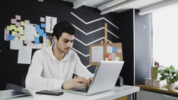 Mann im Büro arbeitet am Computer, schaut sich die Notiz am Telefon an — Stockvideo