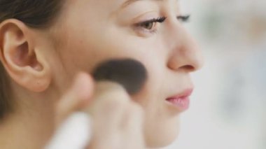 Makyaj yapan bir kadın. Güzel kız aynaya bakıyor ve büyük bir fırçayla kozmetik uyguluyor. Kızın elmacık kemikleri kızarıyor. Pudra, allık..