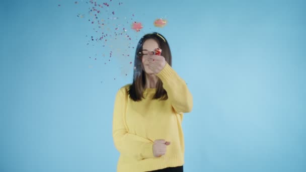 Jeune femme explosant un biscuit confetti sur fond bleu . — Video