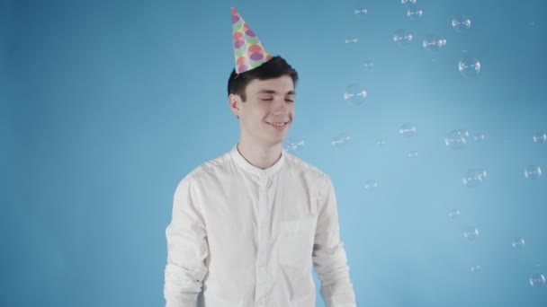 Joven con sombrero de fiesta se sorprende burbujas voladoras sobre un fondo azul . — Vídeo de stock