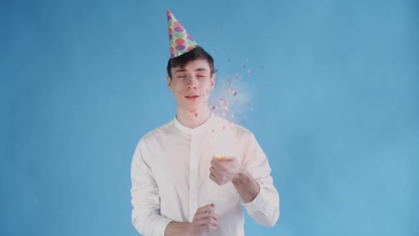 Junger Mann explodiert Konfetti-Cracker auf blauem Hintergrund. — Stockvideo