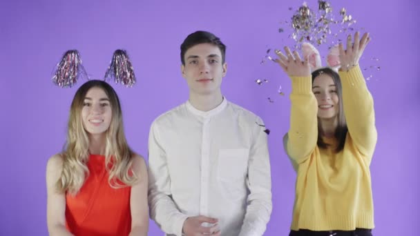 Two beautiful girls throw confetti and young man Exploding party popper on purple background — Stock Video