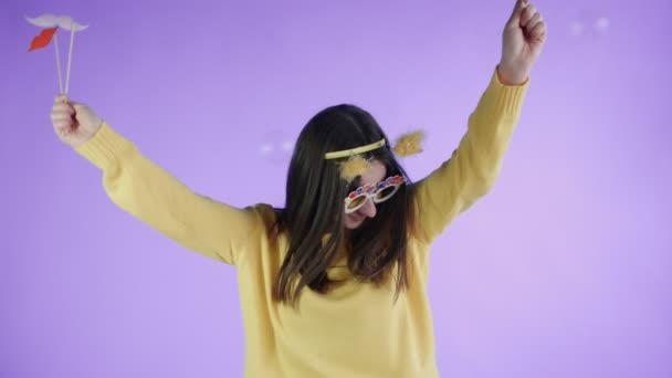 Krásná mladá žena s vtipnou kloboukem a sklenicemi tančí sama na purpurovém pozadí. — Stock video