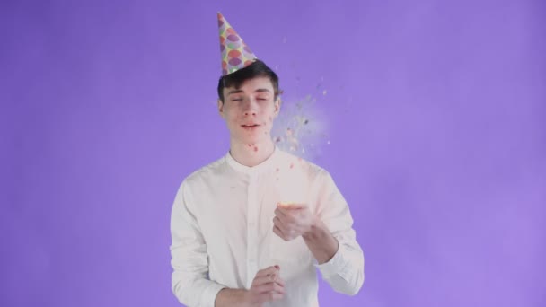Junger Mann explodiert Konfetti-Cracker auf lila Hintergrund. — Stockvideo