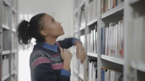 Joven afroamericana mujer está tomando libro de estante en la biblioteca — Vídeo de stock