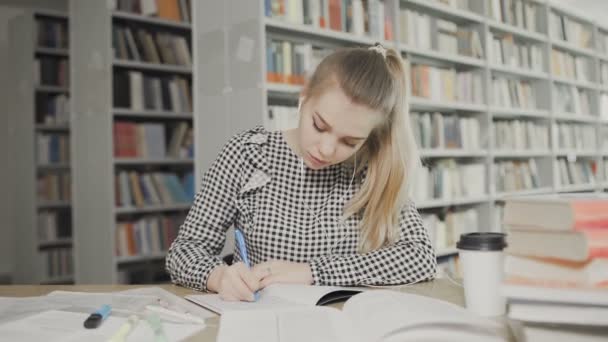 Молодая студентка пишет в своих учебниках в библиотеке — стоковое видео