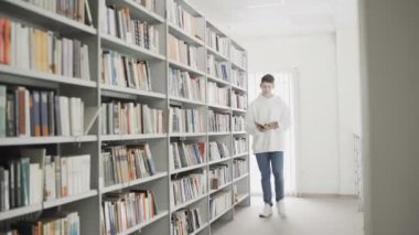 Kitap rafları arasında elinde kitapla dolaşan yakışıklı genç bir öğrenci.
