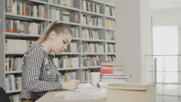 Konsentrert jentestudent med hodetelefoner som forbereder seg til eksamen og skriving mens hun sitter ved universitetsbiblioteket – stockvideo