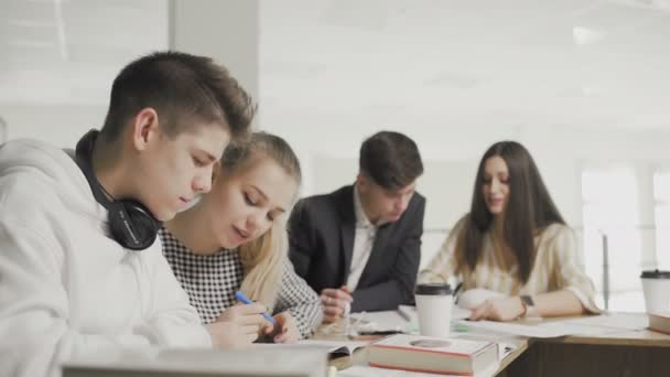 Dos hombres y dos mujeres estudiantes universitarios que estudian con libros y escriben notas en cuadernos en la biblioteca de la escuela secundaria contra la estantería . — Vídeos de Stock