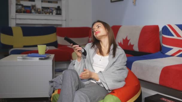 Young woman watching tv and clicking change channels with remote control in hand. Beautiful female changing channel with remote controller while sitting near sofa with cushions with country flags — Stock Video