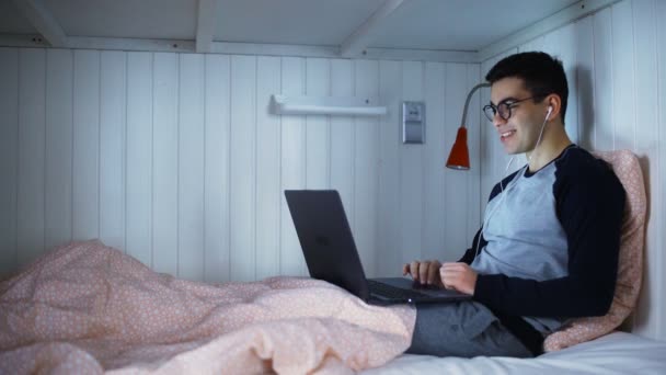 Hombre acostado en una cama y hablando en una cámara web y sonriendo — Vídeos de Stock