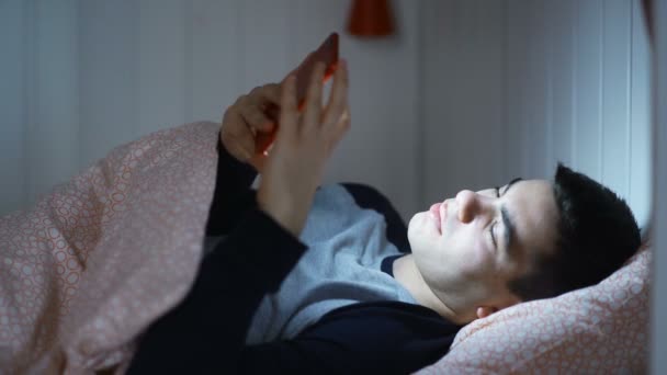 夜遅くに自宅でベッドに横たわっているスマートフォンを使用して若いハンサムなヒップスター男. — ストック動画