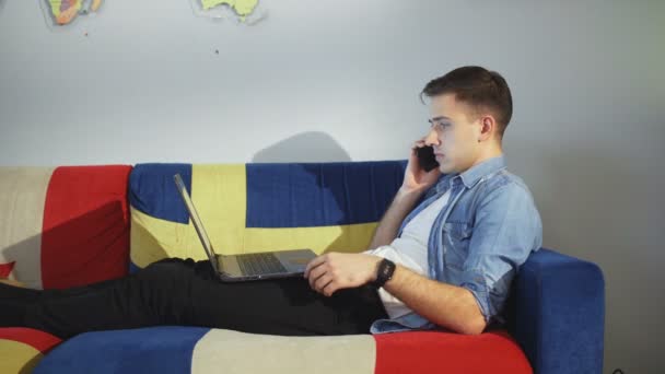 Jóképű boldog ember otthon, leül a kanapéra, miközben füzetet használ és telefonál. — Stock videók