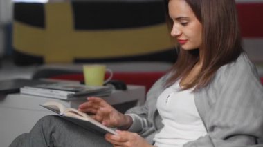 Kanepenin yanındaki yerde kitap okuyan bir kadın. Üzerinde kır bayrakları olan yastıklar var.