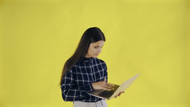 Chica joven que trabaja en el ordenador portátil comprobar las redes sociales en ropa casual y mirando a la computadora en el fondo amarillo en el estudio — Vídeo de stock