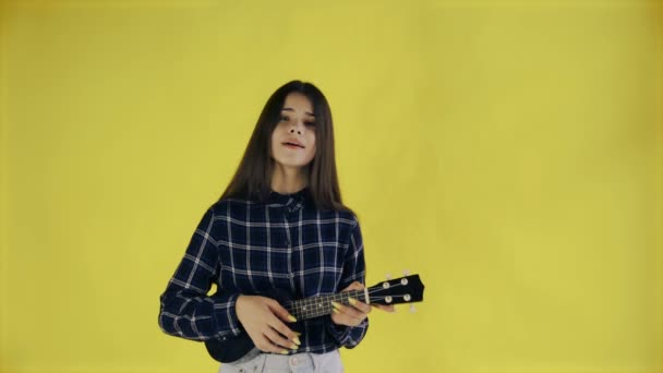 Mladá dívka zpívá a hraje ukulele na žlutém pozadí ve studiu — Stock video