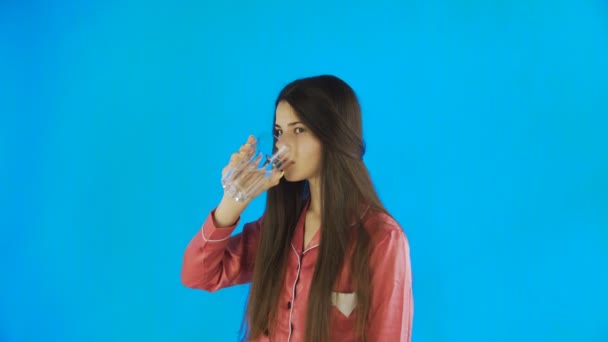 O adolescentă caucaziană bea un pahar cu apă. O tânără bea apă din sticlă pe fundal albastru în studio — Videoclip de stoc