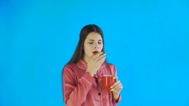 Молода жінка в піжамі п'є чай або каву на синьому фоні в студії — стокове відео