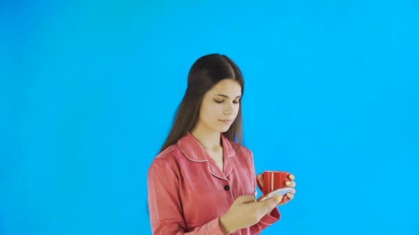 Menina feliz em pijama está bebendo chá ou café e usando telefone no fundo azul em estúdio — Vídeo de Stock