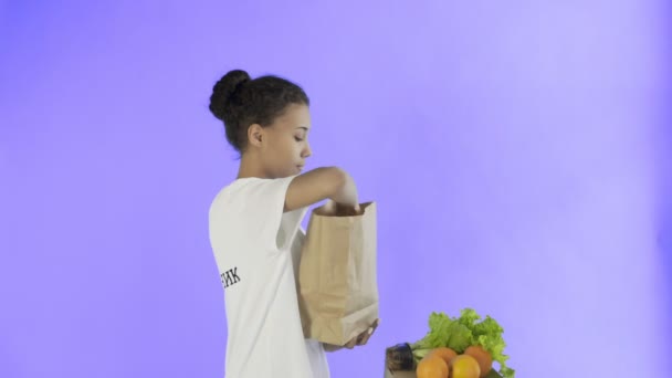 Gelukkig vrouw laden papieren zak met biologische gezonde groenten en fruit op Violet achtergrond — Stockvideo