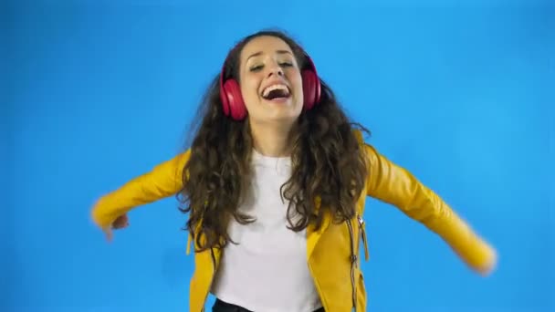 Young woman in yellow jacket dancing while listening music on headphones in Studio with blue Background. — Stock Video