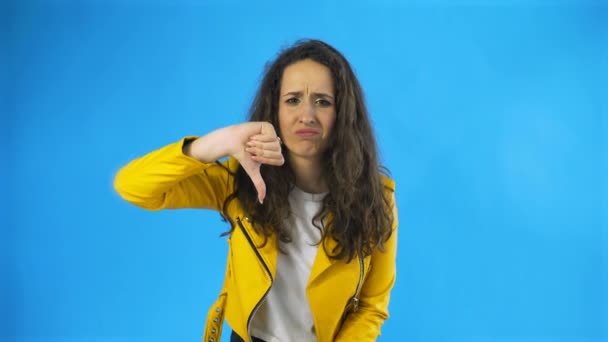 Portrét seriózní ženy ve žluté bundě ukazující palce dolů cedule nelíbí ve studiu s modrým pozadím. — Stock video