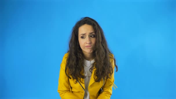 Sad thoughtful young beautiful woman in yellow jacket standing in Studio with blue Background. — Stock Video