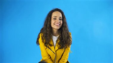 Sarı ceketli genç kadın dudaklarını ısırıyor ve stüdyoda mavi arkaplan ile göz kırpıyor..