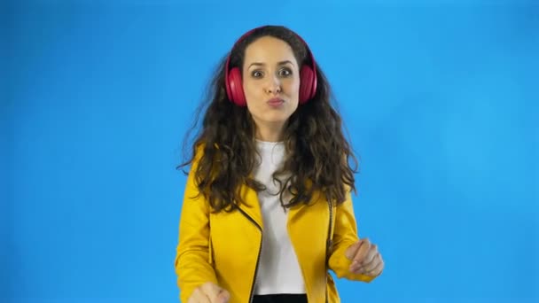 Grappige en leuke vrouw in gele jas is dansen tijdens het luisteren naar muziek op koptelefoon in Studio met blauwe achtergrond. — Stockvideo