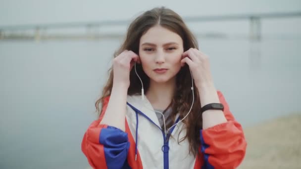 Young woman is lokking at the camera and wearing headphone for music motivation for cardio training, river on background — Stock Video