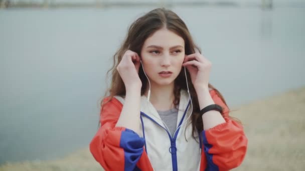 Ung kvinna bär hörlurar för musik motivation för konditionsträning, flod eller hav på bakgrunden — Stockvideo