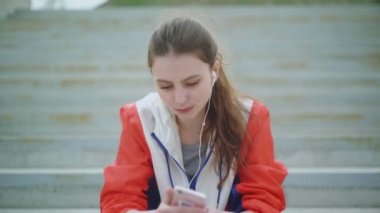 Çekici kadın koşucu merdivenlerde oturuyor, telefon kullanıyor ve yoğun antrenman sonrası yorgun dinleniyor.