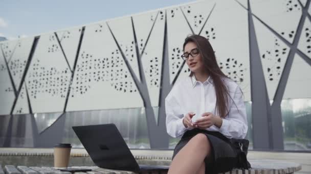 Jovem mulher de negócios caucasiana com longa conversa de cabelo no skype usando laptop sentado ao ar livre . — Vídeo de Stock