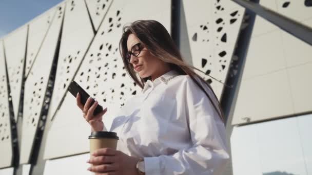 Hermosa mujer de negocios en gafas mirando el teléfono inteligente y beber café. Edificio moderno sobre fondos — Vídeo de stock