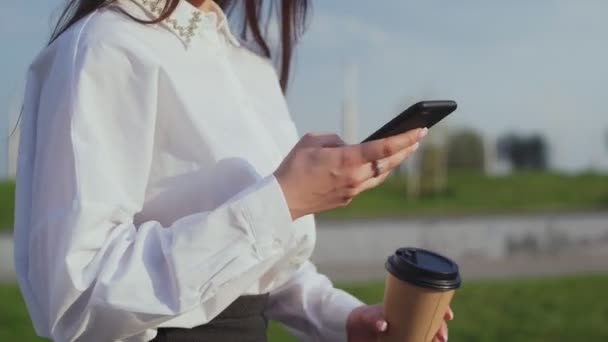 Улыбающаяся элегантная женщина гуляет в городском парке с кофе и смартфоном — стоковое видео