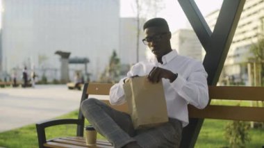 Beyaz gömlekli ve gri pantolonlu genç Afrikalı Amerikalı işadamı parkta yürüyor ve kruvasan yiyor.