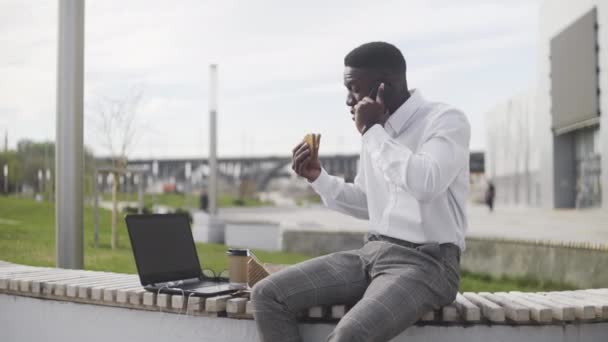 Retrato del joven hombre de negocios afroamericano relajándose y comiendo croissant en el banco en el parque de la ciudad — Vídeo de stock