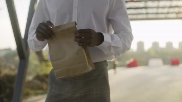 Joven hombre de negocios afroamericano en camisa blanca tomó un croissant del paquete — Vídeos de Stock