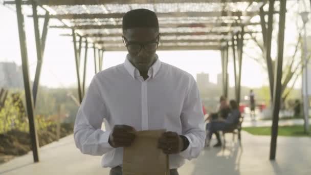 Hombre de negocios afroamericano con camisa blanca bebe café en el parque cerca de la oficina — Vídeos de Stock