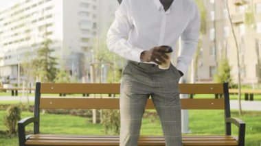 Genç Afrikalı Amerikalı adam akıllı telefonda uygulama kullanıyor ve ofisin yanındaki parkta kahve ya da çay içiyor.