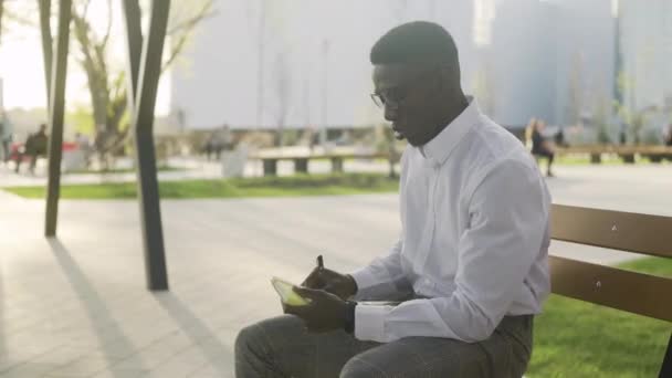 Joven afroamericano en un banco en el parque escribe sus pensamientos en un diario — Vídeo de stock