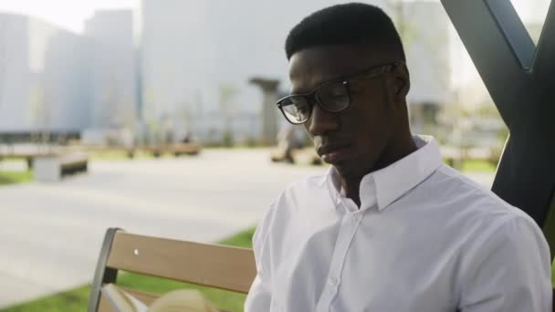 Joven estudiante africano en gafas está sentado en el parque, leyendo libro y tomando café — Vídeo de stock