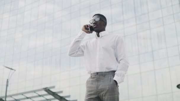 Hombre de negocios afroamericano con gafas hablando por teléfono celular, de pie cerca de su oficina — Vídeo de stock