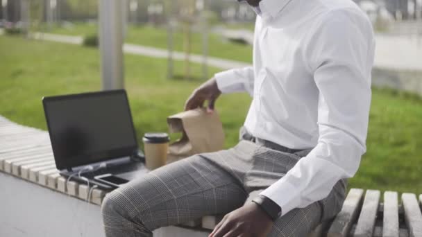 Приваблива афроамериканець людина, використовуючи додаток на смартфон і пити каву або чай в парку біля офісу — стокове відео