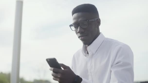 Joven afroamericano usando smartphone y tomando café o té en el parque cerca de la oficina — Vídeo de stock