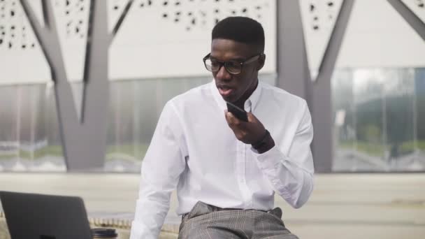 Κομψός Αφρικανός Αμερικανός επιχειρηματίας που καταγράφει ένα φωνητικό μήνυμα ενώ κάθεται στο πάρκο κοντά στο γραφείο — Αρχείο Βίντεο